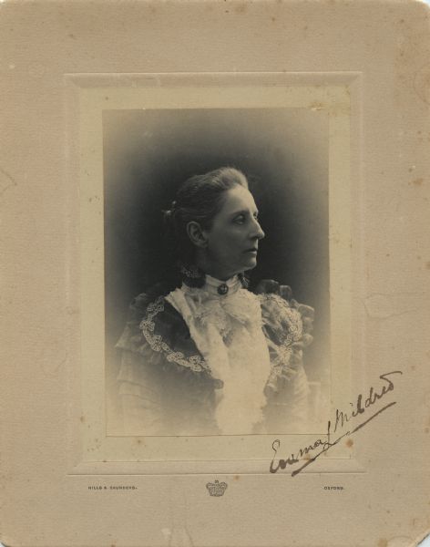 Waist-up studio portrait of a woman sitting in profile. She is wearing a lace blouse with a collar pin, and a dress with elaborate beaded trim. Emma has signed the portrait on the bottom right.