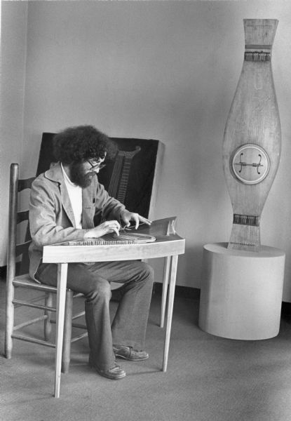 John Pritchard sitting in a chair playing a stringed wooden instrument. There is a large instrument on a base next to him on the right. Pritchard built both instruments.