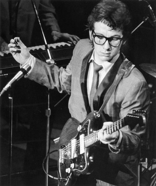 Slightly elevated view of Elvis Costello (Declan MacManus) playing his guitar during a performance. A band member is playing a keyboard behind him.