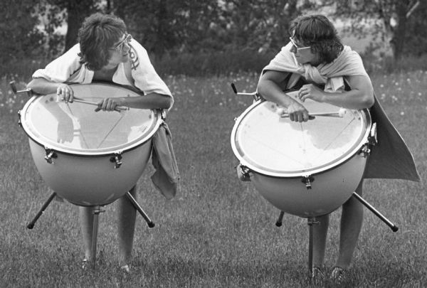Two members of the Madison Scouts Drum and Bugle Corps pausing during a rehearsal outdoor. They are standing and leaning over on their timpani drums.