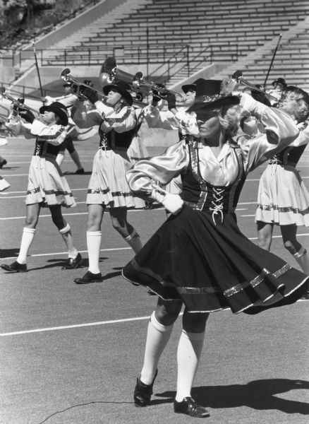 An outdoor performance at a stadium by the CapitolAires, an all-female drum and bugle corps. In the foreground a woman is dancing while buglers are marching behind her.