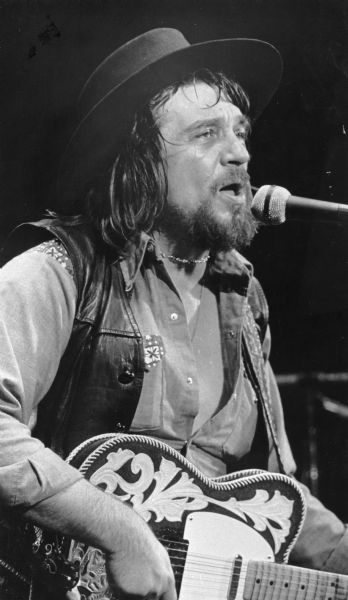 Close-up of musician Waylon Jennings playing guitar and singing during a concert.