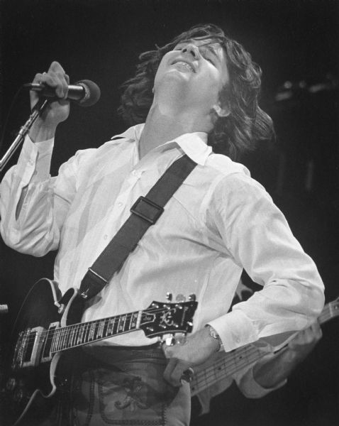 Musician Steve Miller during a concert. His guitar is on a strap around his neck and he is gripping a microphone.