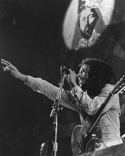 Bob Marley reaching out with one hand, and gripping a microphone in his other hand while performing in concert.