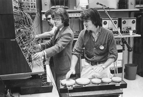 From left to right, Stu Baker and Gregory Patrick Garvey are working at a synthesizer panel, while Serge Ossorguine is playing electronic drums. There are several reel to reel tape decks behind the musicians.