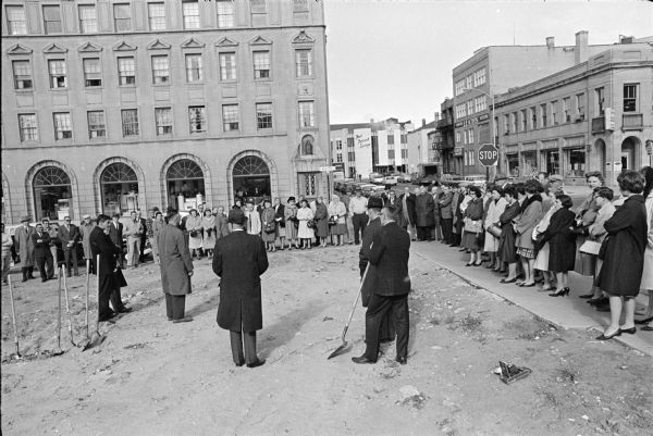 Groundbreaking ceremony for the new Madison Public Library building at the south corner of W. Mifflin and N. Fairchild Streets. Rev. Joseph Brown is standing in the left foreground, Library Director Bernard Schwab is behind Rev. Brown. Kermit Frater is standing center foreground, perhaps speaking. Mayor Henry Reynolds is standing foreground center wearing a hat with his back to the camera. Robert Jones (in a hat) and Robert McMullen are standing with their backs to the camera right foreground. Several shovels are stuck in the ground at left, and a camera is sitting on the ground at right. Several spectators are standing on the sidewalk surrounding the site, and a handful of people can be seen observing from the windows of the Madison Gas and Electric building across the street. Leath's Furniture and First Federal Savings Banks are in the background.