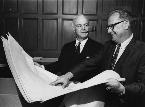 Two men are looking at large papers. One of the men has a pipe in his mouth, and the other is wearing eyeglasses and is smiling and pointing to the sheet of paper. Caption reads: "Officials of Vulcan Materials Co. examine plans for their aluminum processing plant to be constructed in Oak Creek. Bernard A. Monaghan (left) president, and Don H. Warner, president of the aluminum and magnesium division, were here for groundbreaking ceremonies Thursday."