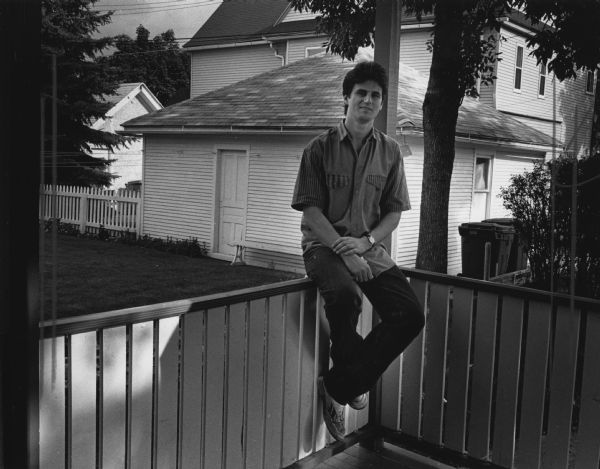 A man is sitting on the railing of a back porch, with a garage and lawn in the background. Caption reads: "Joe Oulahan, 32, came here from Connecticut 15 years ago. He has been a foundry worker at Briggs & Stratton Corp. for 10 years. He and his wife live with their 16-month-old daughter near N. 35th and W. State Streets.