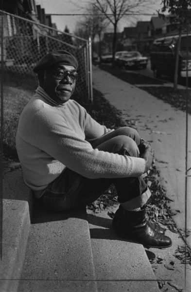 A man us sitting on concrete steps, and is wearing a beret, eyeglasses, and a sweater. Caption reads: "Phillip Moreland, a Milwaukee longshoreman idled by the shutdown of the St. Lawrence Seaway, sat in front of his home at 2872 N. 34th St. Tuesday."