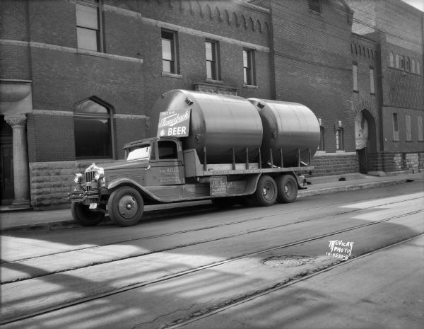 View across street towards two new tanks being delivered by the Heil Co. of Milwaukee on a Heil Company truck. The truck is parked in front of the Fauerbach Brewing Co. at 651 Williamson Street, and the brick building has a stone column and lintel at the corner entrance on the left. A sign above the truck cab reads: "Since 1848 Fauerbach Beer." There are railroad tracks in the street.