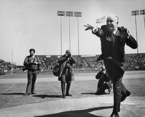 A man wearing a suit has just tossed a baseball. Three men are standing behind him taking photographs. Caption reads: "County Executive John Doyne showed his pitching skill in pregame ceremonies as the Brewers opened the baseball season Tuesday at County Stadium."