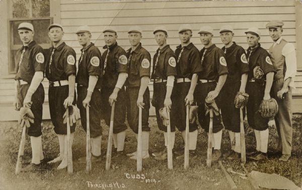 Eleven men standing together, facing left and looking towards the photographer. Ten of the men are wearing baseball uniforms, and nine of them are holding baseball bats with their left hands. The man on the far right is wearing a vest, trousers and a cap. There is a building behind the group.