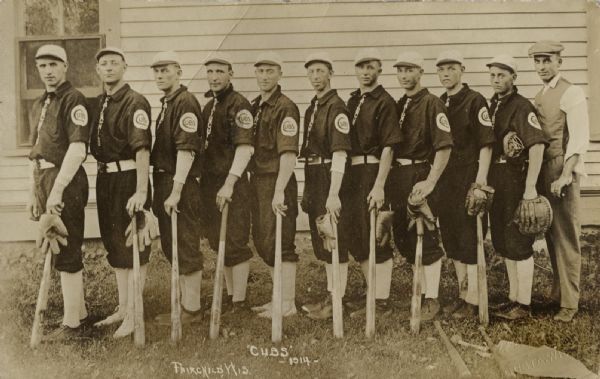 Eleven men standing together, facing left and looking towards the photographer. Ten of the men are wearing baseball uniforms, and nine of them are holding baseball bats with their left hands. The man on the far right is wearing a vest, trousers and a cap. There is a building behind the group. Caption reads: "'Cubs' -1914-" and "Fairchild, Wis."