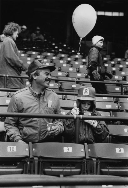 A man is smiling and sitting next to a girl, who is holding a balloon, among the seats in a stadium. They are both holding cups in their hands. Caption reads: "Kevin Kramasz brought his daughter Jessica, 9, to the Brewers' open house Saturday."