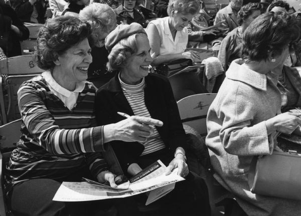 Two women smiling and looking towards where one of them is pointing. They are holding a large flyer (a scorecard?) and are sitting among other women in a stadium. Caption reads: "<b>Sisters can enjoy baseball,</b> even when only one is a "pro." Mrs. Louis Kische (left), 3915 W. Roosevelt Dr., explained a play to her sister, Mrs. Eileen Roloff, 4077 N. 60th St., who was attending her second major league game. The two were among those attending the ladies' day baseball seminar before the Brewers game Thursday at County Stadium."