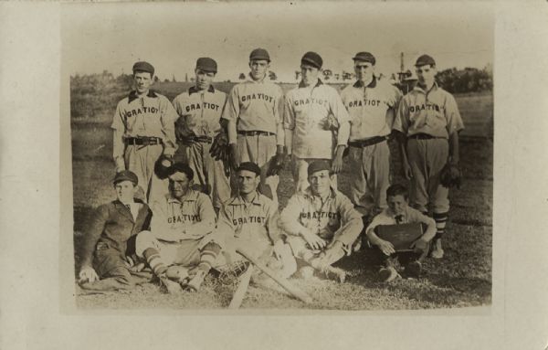Outdoor group portrait of ten men and a boy. Nine of the men are wearing baseball uniforms, and the man on the far left is wearing a coat and hat, and the boy on the far right is wearing a shirt and necktie. The uniforms read "Gratiot."