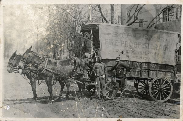 George William Harris is standing on the left at the side of a mule-driven moving van. Another man is standing next to him on the right. The van is parked on an unpaved street, and trees and houses are in the background. The moving company was Stacey's Fire-proof Storage, based in Cincinnati, Ohio at 2333-2339 Gilbert Avenue.
