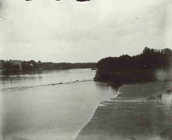 View along shoreline of the Wisconsin River, with a sandy beach leading to a stand of trees on the bank on the right. Buildings are along the opposite bank.