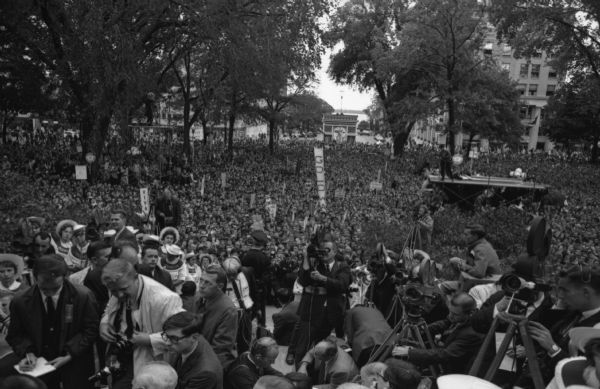 Large crowd gathered at the Wisconsin State Capitol to hear Senator Barry Goldwater speak. Reporters and photographers are gathered in the foreground. A miniature Arc de Triomphe can be seen in the background at the Monona Avenue entrance to the Capitol (now Martin Luther King Jr. Blvd.).