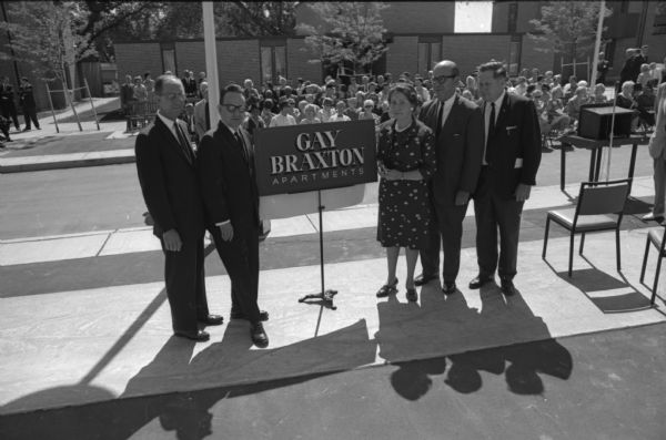 Guests honored at the dedication ceremony of the Gay Braxton Apartments gathered around a sign for the building. They are left to right Mayor Otto Festge, former MHA chair Roland Day, Mary Lee Griggs, colleague of the late Neighborhood House director Gay Braxton, federal Public Housing Authority official P.F. Papadopulos, and current MHA chair realtor Earl Espeseth. A group of new residents is in the background.