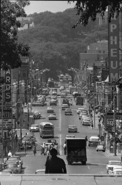 View down State Street from the Capitol Square towards Bascom Hill. Cars and pedestrians are along the street, which is lined with signs and businesses.