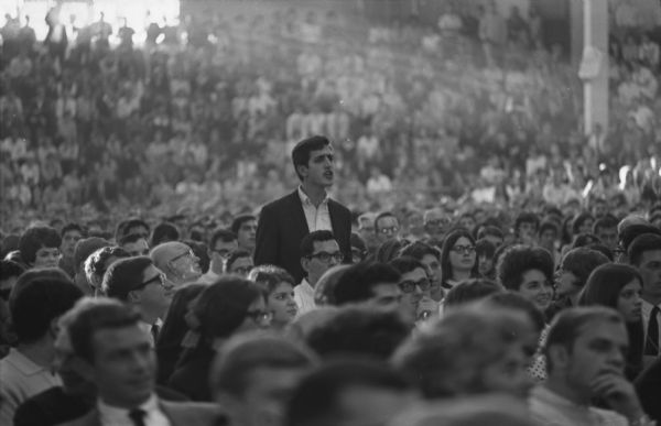 View across crowd of people towards a man standing. The crowd is assembled to hear Ted Kennedy speak at the University of Wisconsin-Madison Stock Pavilion.