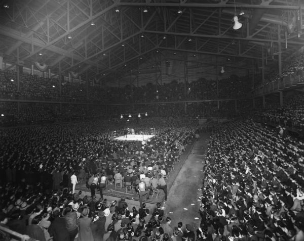 Elevated view of a full crowd at the Field House, including people sitting on the floor in chairs watching the boxers in a boxing ring in the center of the floor.