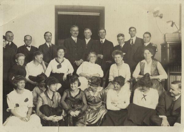 A group of men are posing standing behind two rows of women sitting. The men are all wearing suits, and the women are wearing a variety of outfits. One woman's dress has two paper jack-o-lanterns affixed to it.