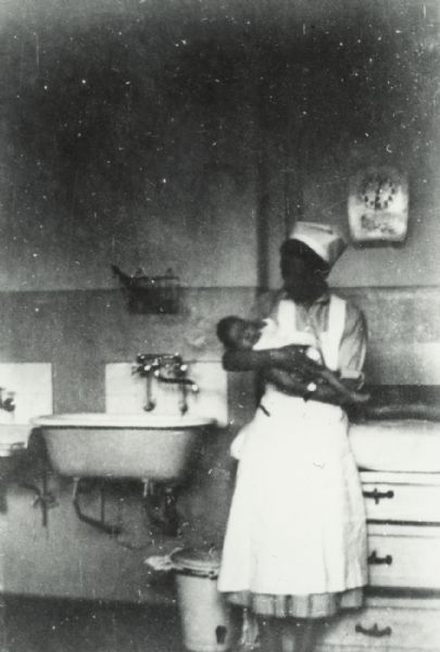 A woman in a nurse's uniform is standing and leaning against a cabinet while holding an infant in her arms. Behind her is a clock, a sink, and a wastebasket. 