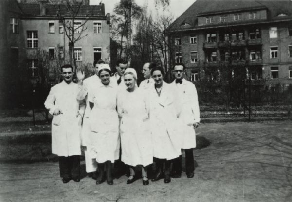 Group portrait of eight men and women in lab coats, standing outdoors, with two buildings in the background. Caption reads: "Israelitisches Krankenhaus was a teaching hospital of 350-500 beds, completely kosher food, Jewish Sabbath and holidays observed. Medical staff was exclusively Jewish; patients not. Hospital destroyed ca. 1938."