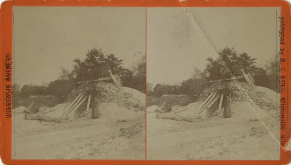Stereograph of a woman standing outside a structure made from slabs of rock and wooden support beams. Caption reads: "Lime kilns. 'Old La Fever' 3 miles S. of New London."