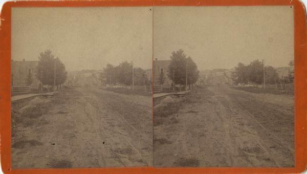 Stereograph view down an unpaved road, with trees and buildings on either side. A large white house is faintly visible at the end of the road. Caption reads: "View of So. Main Str. (white house) Dittberner tailor shop at the corner 8th & So. Main. Taken 1894 from corner 13th & No. Main, present location of Cam Hosp- [?] on left hand side of road."