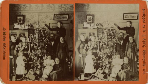 Stereograph view of eight people posing around a Christmas tree. Caption reads: "M. Dittberner & family, B.G. Stieg & family, Anna & Mary Block."
