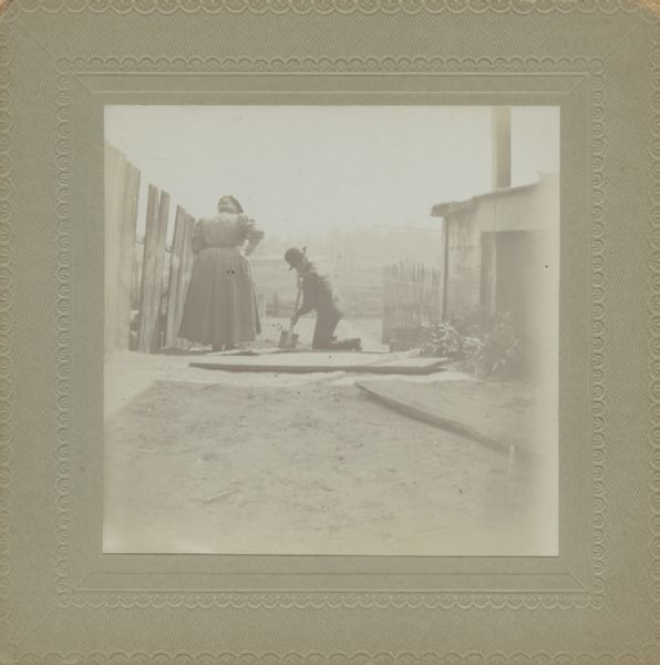 A man and a woman are standing outside between a fence and a building. The man is kneeling and using a shovel, and the woman is standing and watching him. Caption reads: "Mr. & Mrs. Wm. Dittberner gardening, 75 So. Main."