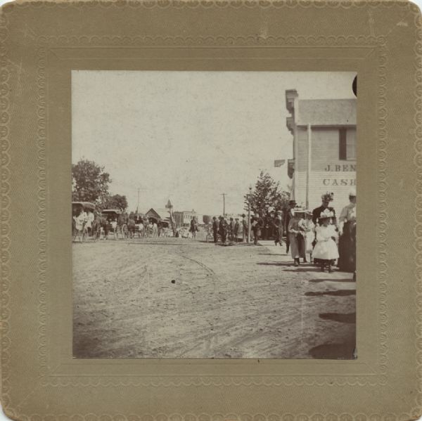 View down unpaved road through town, with a parade of horse-drawn wagons on the left, and people walking along a sidewalk on the right. A building on the right has a sign painted on the side. Caption reads: "Decoration Day parade, corner of 7th & So. Main."