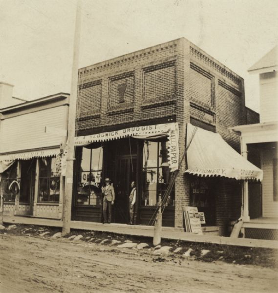 View from unpaved street towards two men posing in front of a brick building, which has signs that read: "F. A. Sedgwick Druggist," "Wallpaper," and "Stationery." Caption reads: "Mr. Kaiser, pharmacist, Mr. F.A. Sedgwick, druggist."