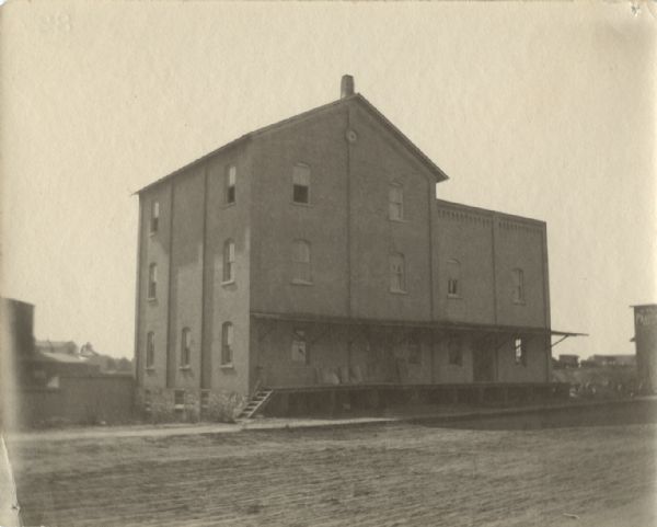 Exterior view across unpaved road towards a three-story building with a loading dock in front. Many of the windows are open. Caption reads: "Hoffmans feed mill, 1904."