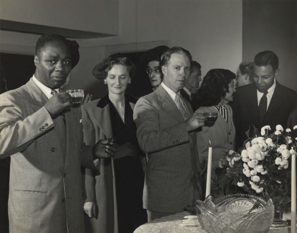 Six people stand near a table with a punch bowl, some posing for the camera and others seeming unaware of it. Caption reads: "Reception for Canada Lee, stage and screen actor, held at Y.W.C.A. October 14, 1945, by the Madison branch of the National Association for the Advancement of Colored People. Left to right: Canada Lee, Mrs. Karl Link [Elizabeth Feldman], Miss Jane Lester, Melburn Heinig, violinist, Mrs. Sanford Farness [Mary Jane Miltgen], and Ted Pierce."