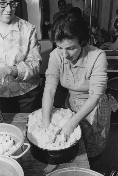 A woman is pressing her hands into dough in a large pot while another woman is looking on. Other containers are on the table, and at least one includes chopped food. Other people are in the background.