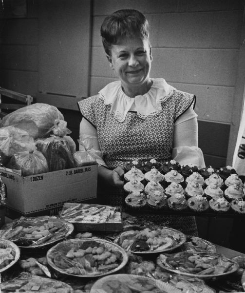 A woman is smiling and is holding up a tray of decorated cupcakes. In front of her is a table holding several wrapped plates of cookies and a box with bagged bread. Caption reads: "<b>Displaying cupcakes</b> she made and decorated with Christmas colors, Mrs. Bert R. Krahn [Marcella E. Maas], West Bend, took a turn at selling in the Hillside Homemakers' club booth at the annual fall fair, Richfield. Plates and jars filled with cookies, caramel apples, loaves of bread and caramel popcorn as well as craft items were sold."