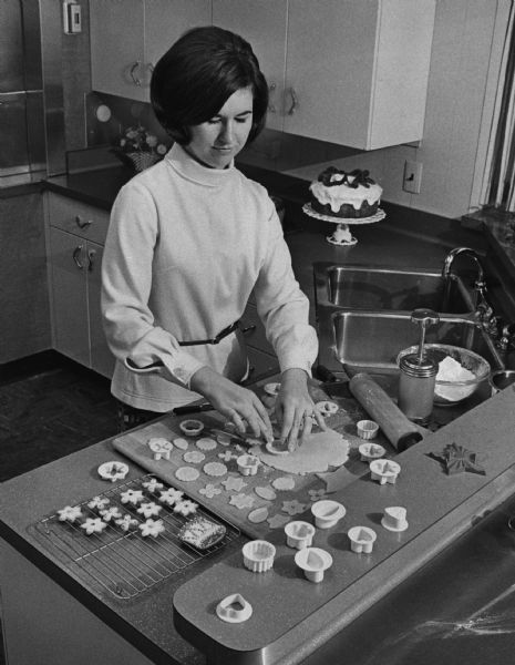 A woman is pressing a cookie cutter into rolled dough at a kitchen counter. Around her are cookie cutters, a rolling pin, and cookies on a cooling rack. A decorated cake is in the background. Caption reads: "MISS KAY BOSSMAN, 1106 E. Knapp St., made spritz cookies in delicate flower shapes. She is home service adviser for the Wisconsin Gas Co. kitchens."