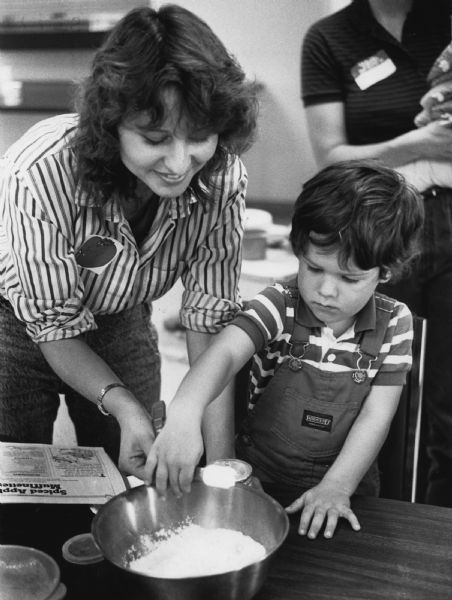 A woman is helping a boy pour an ingredient from a measuring spoon into a mixing bowl. Next to the bowl is a recipe for Spiced Apple Muffinettes. Caption reads: "Suzy Michel helped Nicholas Appleby, 3, as he measured flour for the spiced apple muffins the children were baking."
