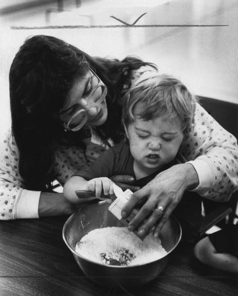 A woman is holding a boy who is scooping an ingredient out of a container into a mixing bowl. Caption reads: "Fran Kozel and her son, Samuel, 2, measured cinnamon (above) during a cooking class Monday for children at the Logemann Community Center in Mequon."