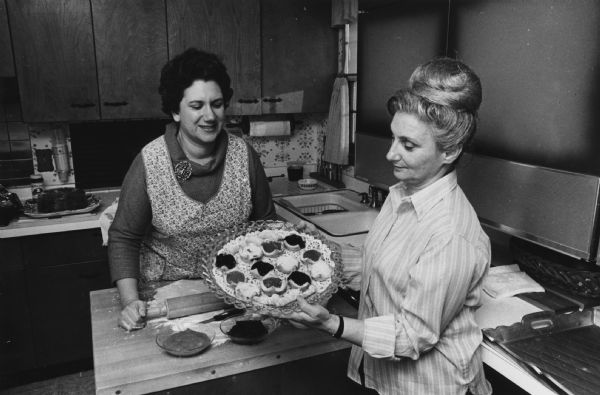 Two women are standing in a kitchen. One woman is standing behind a butcher block counter rolling out dough with a rolling pin, along with bowls of ingredients on it. The other woman is standing in front of the counter and is holding up a plate with completed pastries on it. Caption reads: "<b>Tantalizing odors</b> are filling the kitchens of members of the sisterhood of Temple Emanu-El B'ne Jeshurun these days as the women turn out traditional Jewish pastries and cakes which will be sold at their antiques show Nov. 29, 30 and Dec. 1. Mrs. Morton Kesselman [Florence Slavick] (left), 7415 N. Lombardy Rd., and Mrs. Jack Stern [Lydia Klein], 8453 N. Indian Creek Pkwy., both of Fox Point, joined forces for an afternoon of baking. The show will be held at the temple, 2419 E. Kenwood Blvd."