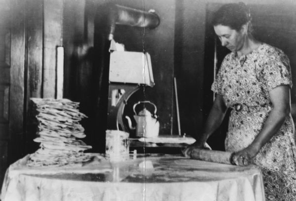 A woman is rolling dough with a rolling pin while standing at a table. On the opposite end of the table are several stacked flat lefse. Caption reads: "Mrs. Odell Oakland [Clara Olson], making lefse. Stepmother of photographer, Lucinda Morken. Prob. North Beaver Creek Valley, Wis."