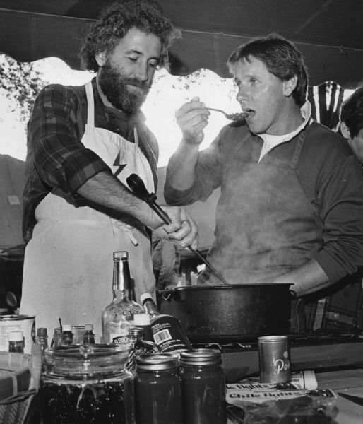 A man is stirring food in a pot, while another man is trying a spoonful of it. A variety of bottles and cans are on the table near the pot. A connected news article provides context: "<b>Hot time at chili cook-off</b> Chili lovers from across the state gathered in Green Lake over the weekend for the 6th Annual Wisconsin State Chili Cook-Off Championship. About 40 teams participated in the event, which was held at Deacon Mills Park. Each team's chili was prepared and cooked at the site on the day of the cook-off and then judged (<b>below</b>). Visitors were able to see if they agreed with the experts as they sampled the chili after the judging. David Fenner, 41, of Janesville, was the winner and will go to California to compete with winners from other states for the national title."  Caption for this image reads: Fred Derr stirred his chili while Dan Buretta sampled it.