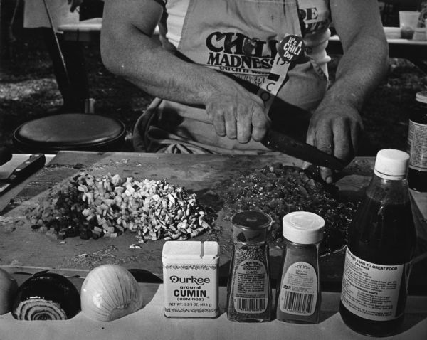 A person (out of frame) is chopping tomatoes on a cutting board, next to a pile of chopped onions. In front of the board are onion halves and containers of ground cumin, rosemary leaves, ground bay leaves, and a bottle of unidentified sauce. The person is wearing a shirt on which the words Chili Madness are visible, and a button that reads "It's A CHILI Day" is affixed to the apron. A connected news article provides context: "<b>Hot time at chili cook-off</b> Chili lovers from across the state gathered in Green Lake over the weekend for the 6th Annual Wisconsin State Chili Cook-Off Championship. About 40 teams participated in the event, which was held at Deacon Mills Park. Each team's chili was prepared and cooked at the site on the day of the cook-off and then judged (<b>below</b>). Visitors were able to see if they agreed with the expert as they sampled the chili after the judging. David Fenner, 41, of Janesville, was the winner and will go to California to compete with winners from other states for the national title."  Caption for this photograph reads: "The ingredients for a good bowl of hot chili were laid out as they were being prepared for the competition."