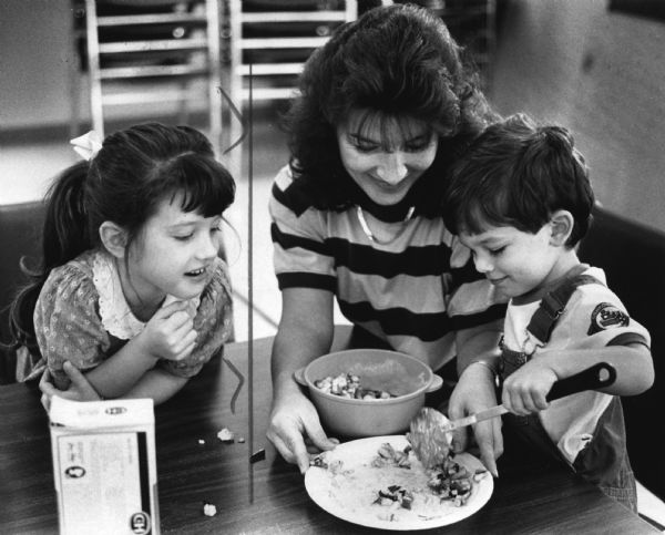 A woman is smiling and holding a plate, while a young boy on the right is ladling food out of a bowl onto the plate. A young girl is watching from the left. Caption reads: "Lucille Nagle held the plate while Thomas, 19 months, handled the spoon."