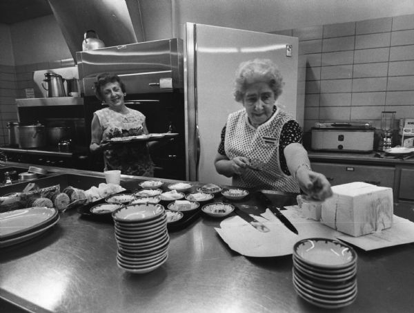 Two women are working in a large industrial kitchen. One woman is holding a tray and smiling, and the other woman is reaching for bowls while holding a spatula. A tray of prepared dishes is on the table between them. Caption reads: "Finding recreation in the kitchen, Miss Fritzie Leiser, 3128 W. Wisconsin Ave., and Mrs. Helen Wickersham, 925 Glenview Ave., Wauwatosa, worked at the senior center of Our Savior's Lutheran Church, 3022 W. Wisconsin Ave."