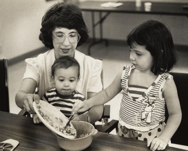 A woman is holding a plate over a bowl while a girl is scraping food from it into the bowl. A small child is sitting on the woman's lap. Caption reads: "Jodi Colosimo held P.J., 2, while daughter Nicki, 4, transferred some chopped apples."